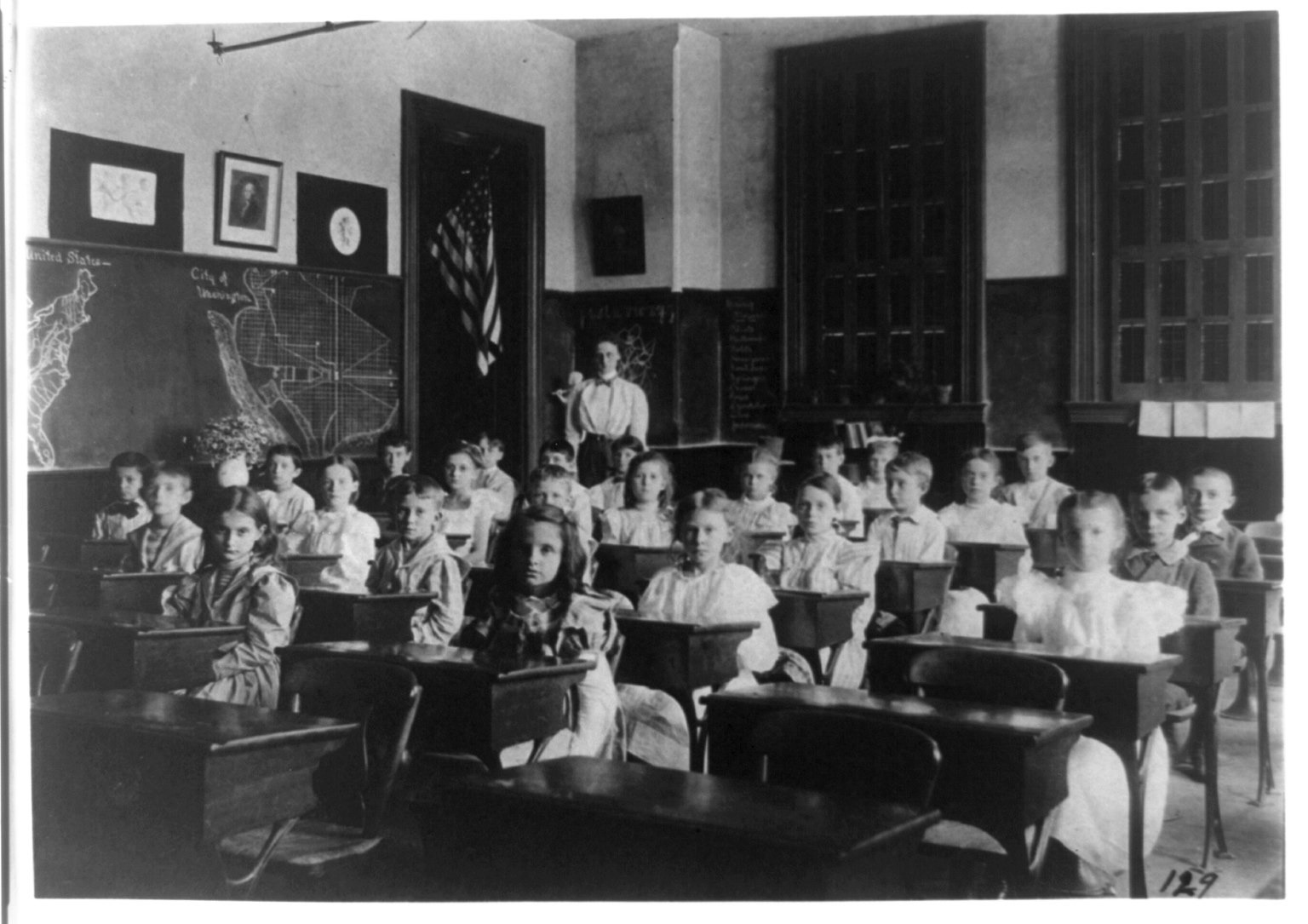 Rows of children in white dresses and sailor suits sit at wooden desks, the teacher standing at the back by a flag, chalkboard maps of the United States and Washington on the wall