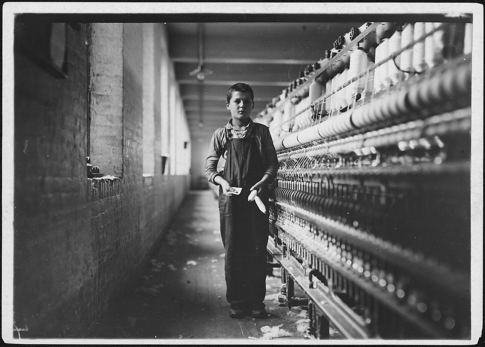 A boy in overalls stands in a narrow mill aisle holding a bobbin, beside a long frame of spinning yarn