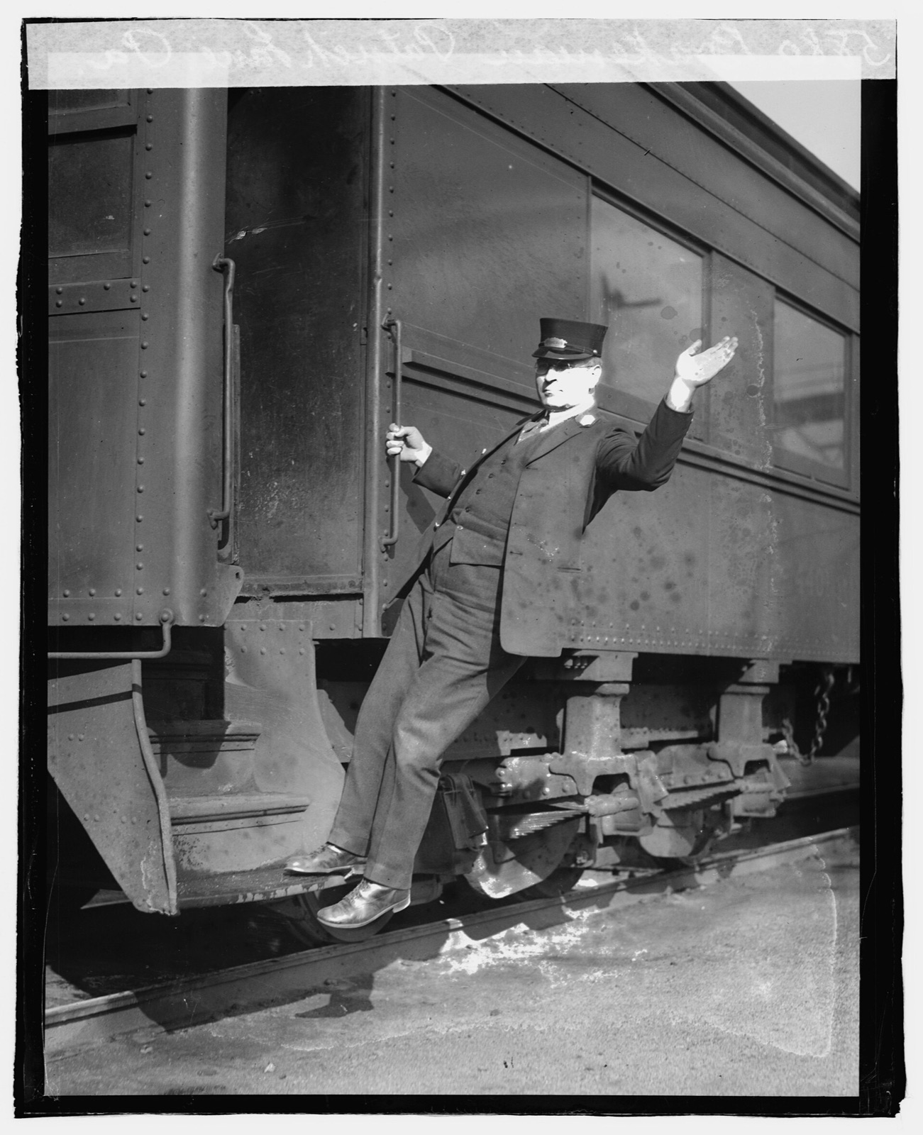 A man in a uniform cap and glasses hangs from the grab iron on the step of a railroad car with one arm raised in a signal