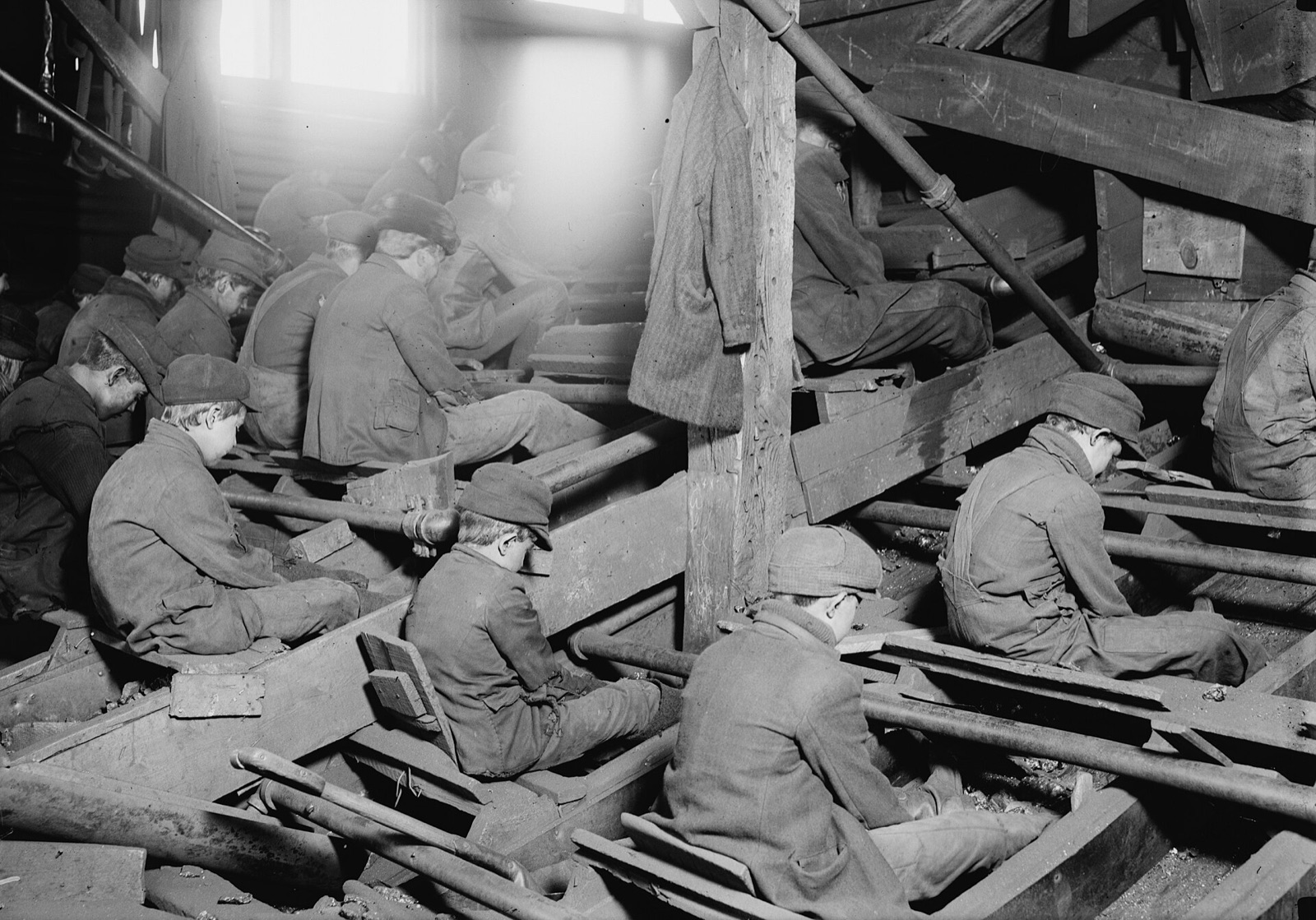 Rows of boys in caps and coats sit hunched over wooden chutes in a dusty coal breaker, picking stones out of the coal running beneath them