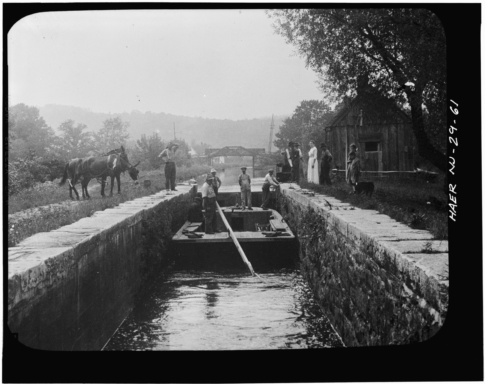 A flat canal boat with men aboard sits low in a stone lock, a man pushing a long pole, two mules waiting on the towpath at left