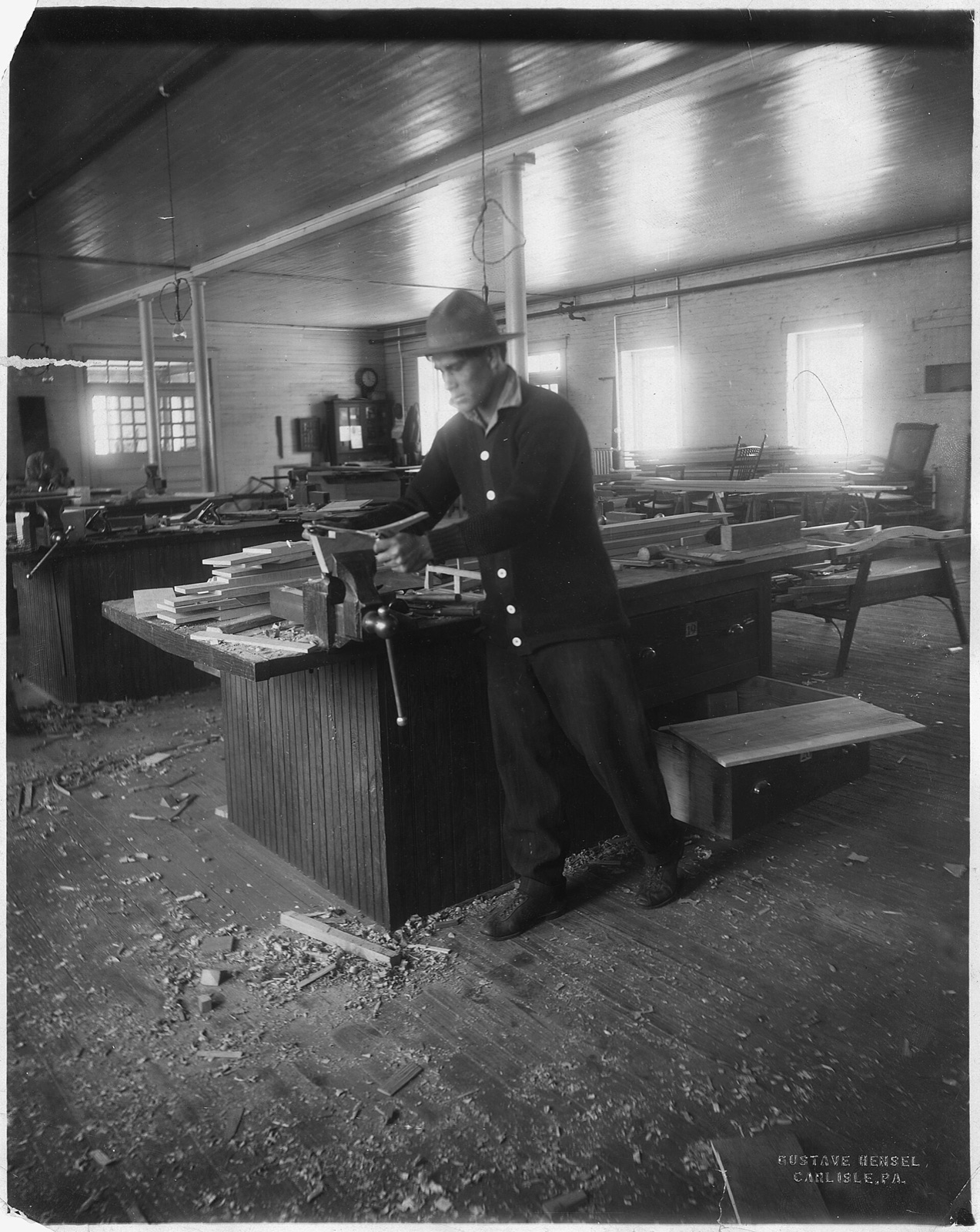 A young man in a hat and cardigan draws a spokeshave along a curved piece of wood held in a vise, in a big workshop strewn with shavings