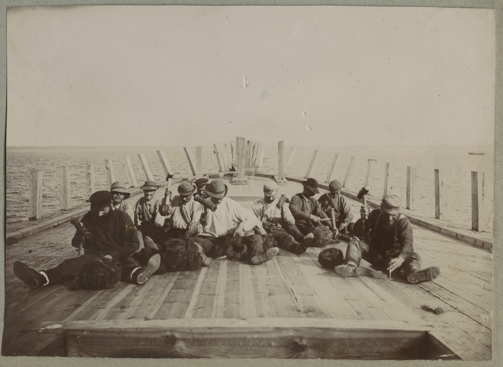 A row of men sitting on a bare wooden deck, swinging mallets to drive tarred oakum into the seams between the planks