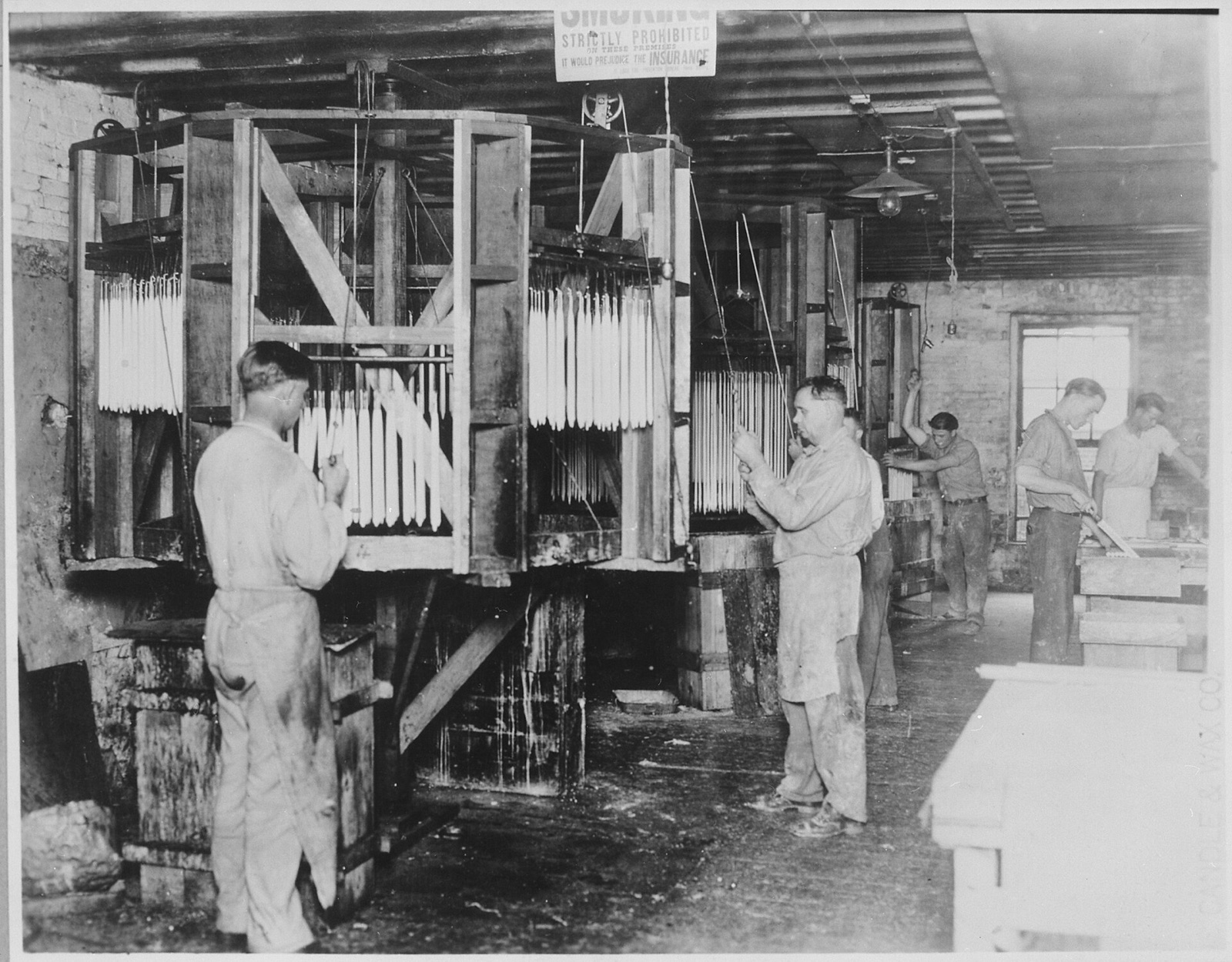 Men in aprons at tall wooden frames hung with rows of white candles being dipped in vats of wax