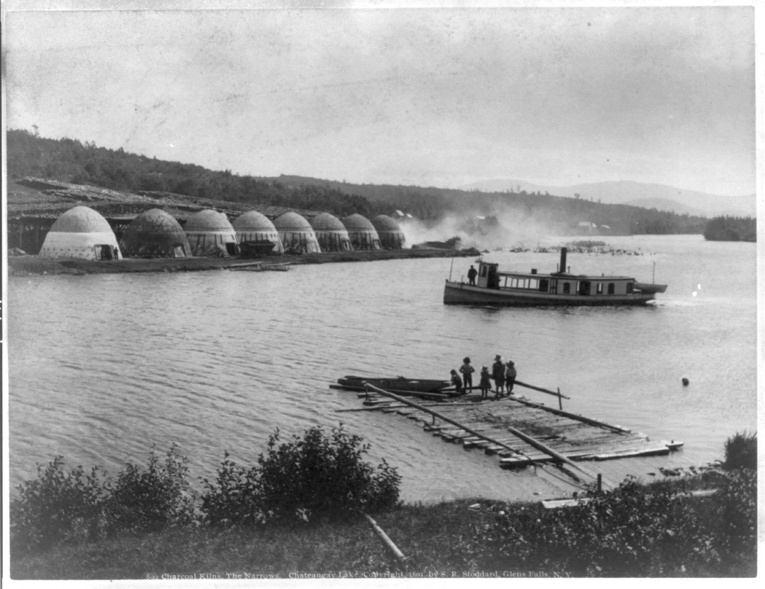 A row of beehive-shaped charcoal kilns along a lake shore with smoke drifting beyond, a small steamer and a log raft with children in front
