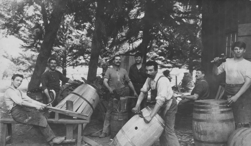 Coopers in shop aprons working outdoors under trees, one shaving a barrel stave on a wooden bench while others drive hoops onto barrels