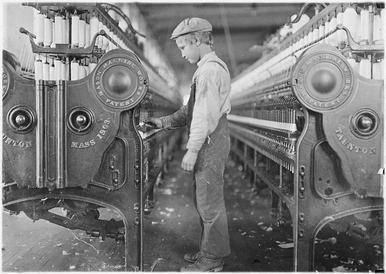 A boy in overalls and a cap reaches into a long spinning frame lined with white bobbins of thread