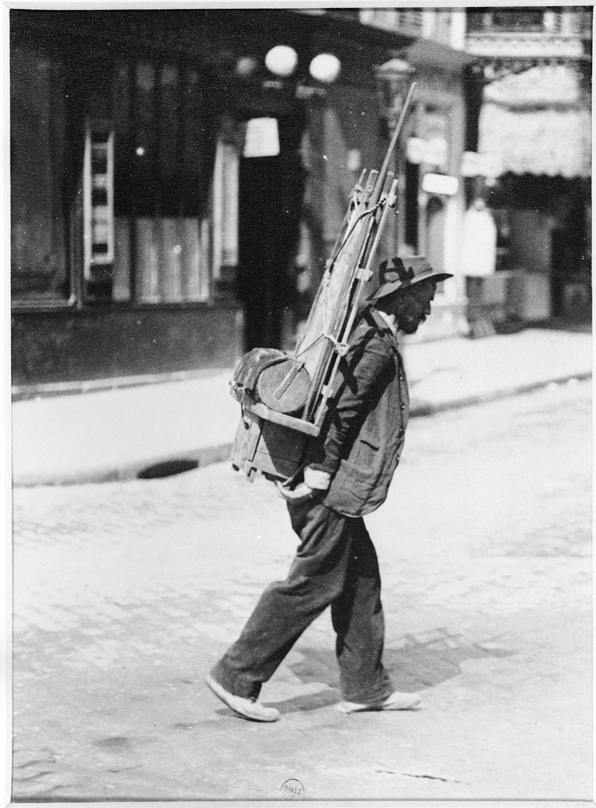 A bearded man in a hat walks across a cobbled street carrying a wooden back rack loaded with sheets of glass and a tool box