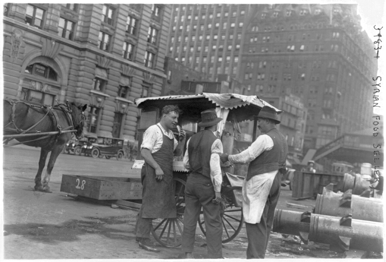 Three men in aprons and vests stand at a covered two-wheeled pushcart on a city street, a horse and tall buildings behind