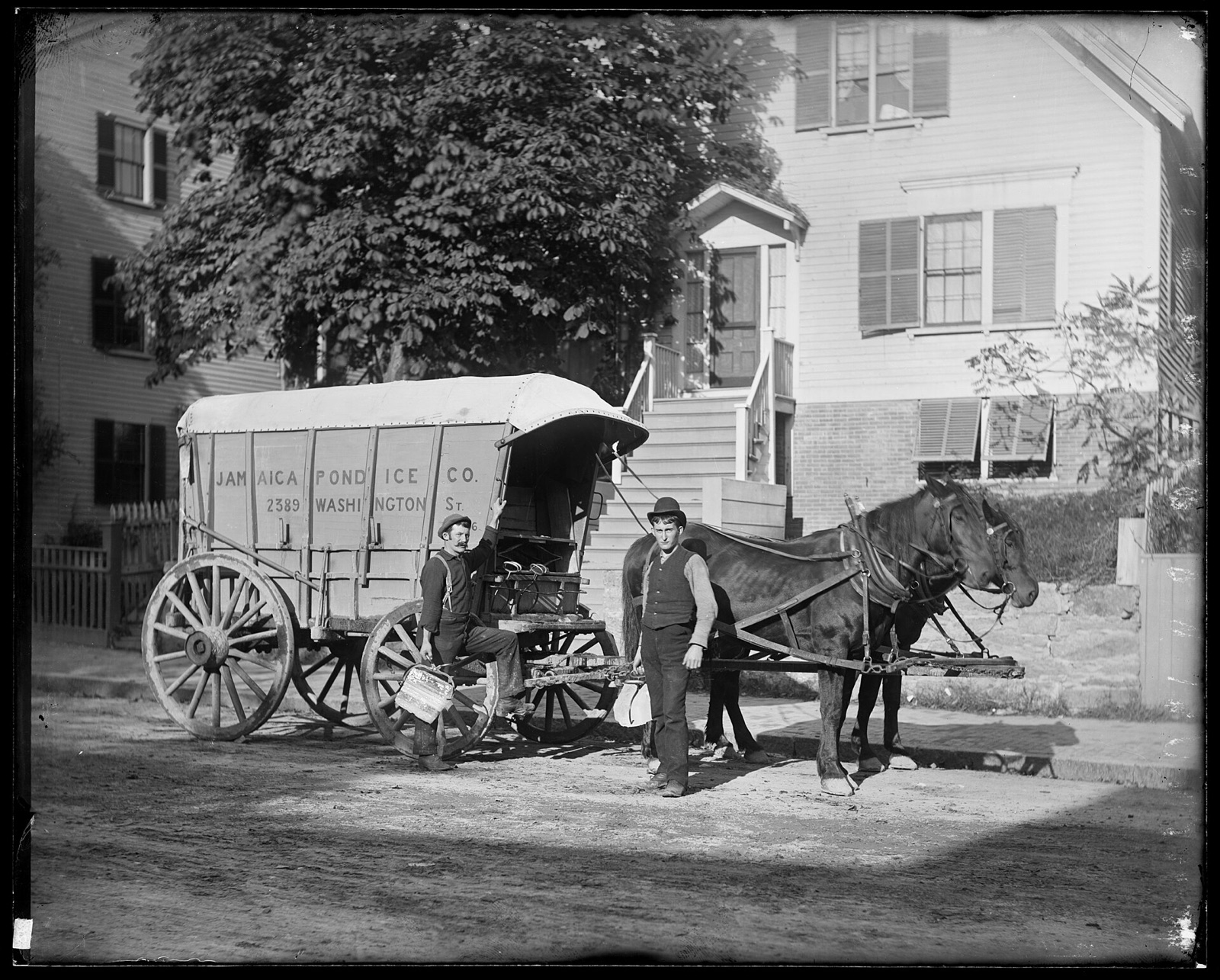 Two men stand by a covered horse-drawn wagon lettered Jamaica Pond Ice Co., one holding ice tongs and a pail, in front of a frame house