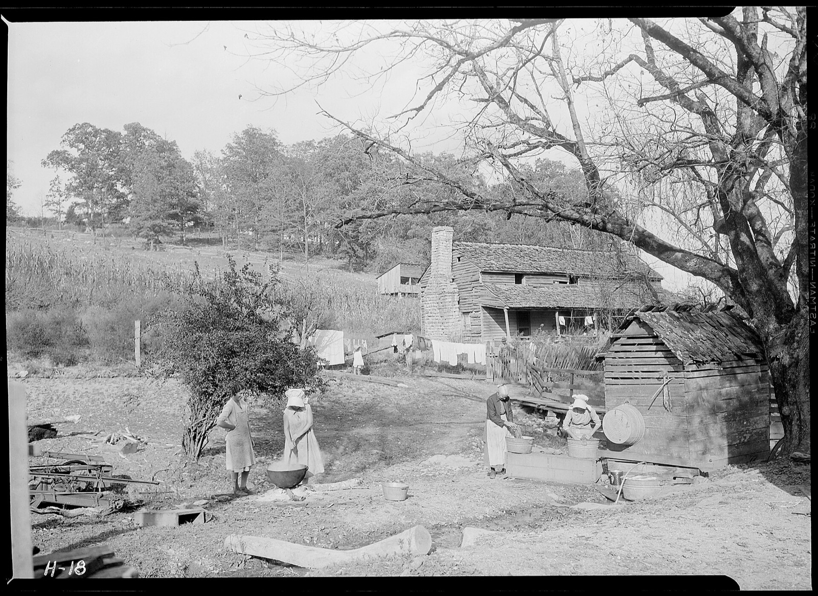 Farm yard with women in aprons and bonnets at washtubs and a big iron kettle, laundry drying on lines before an old farmhouse with a stone chimney