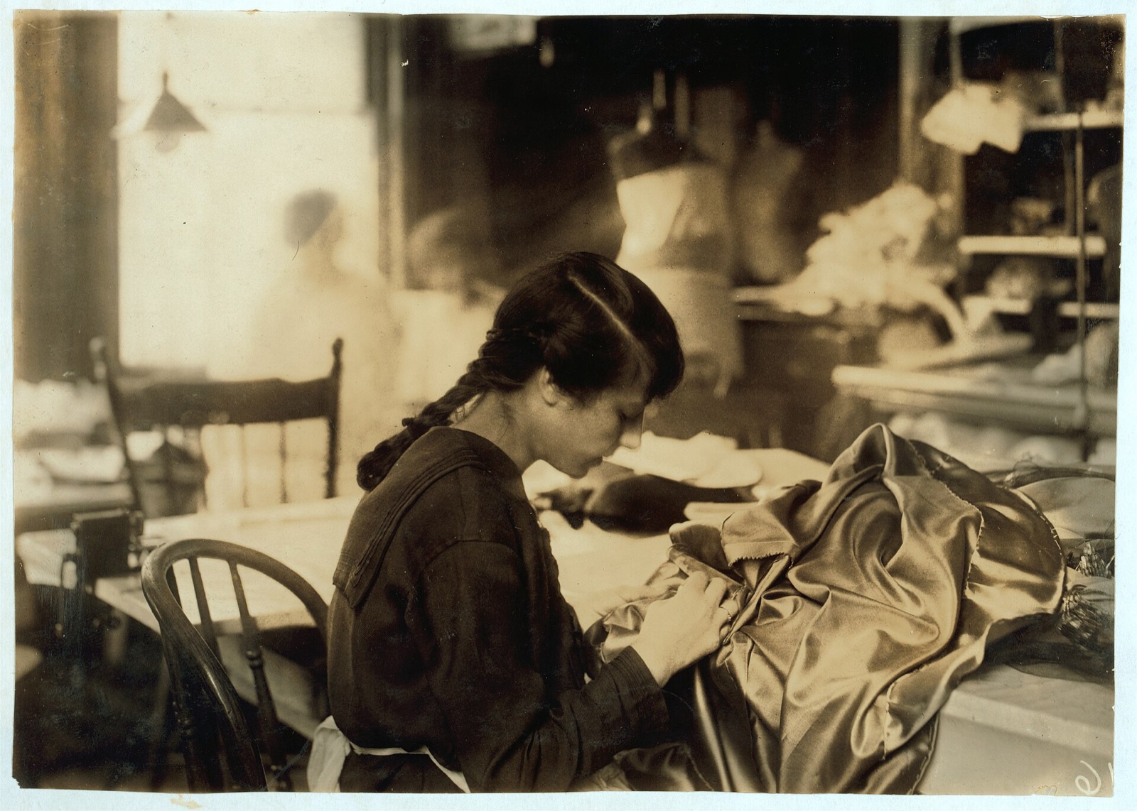 A girl with a long braid bent over a table, stitching shiny satin cloth, with dress forms in the workroom behind her
