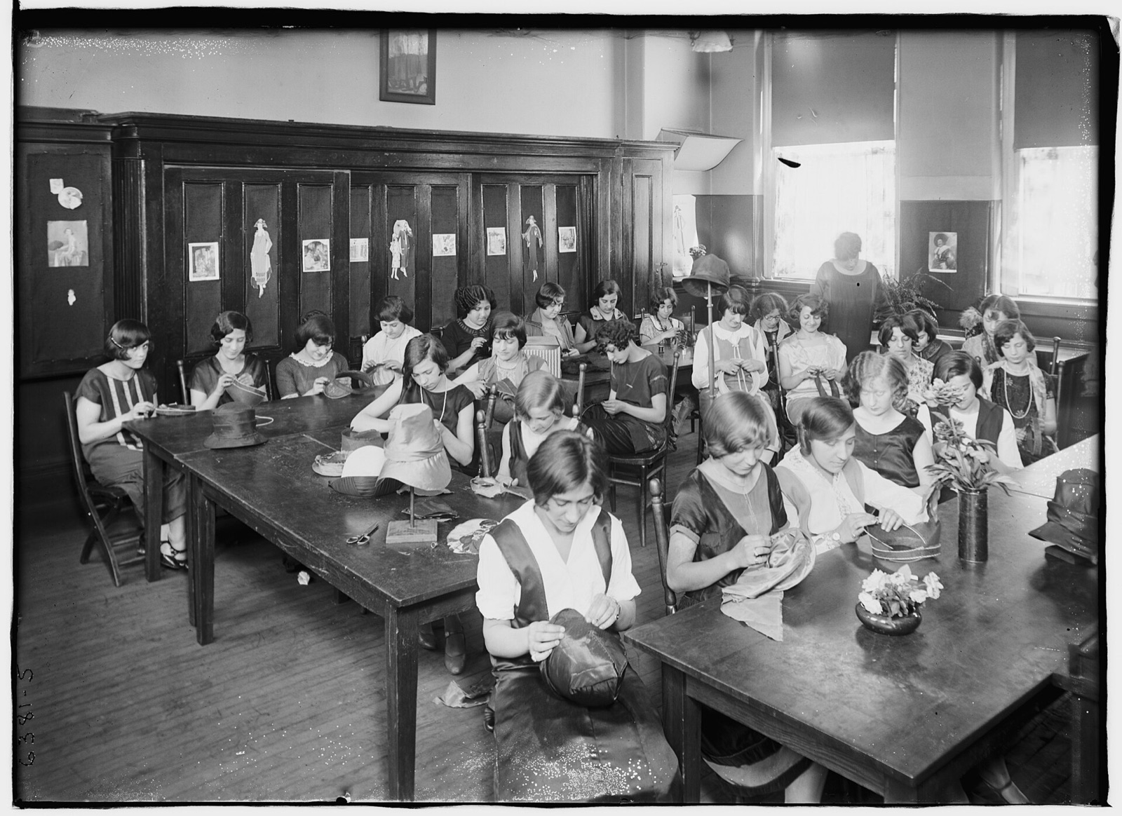 A classroom of young women at long tables sewing hat crowns and brims, with finished hats on stands