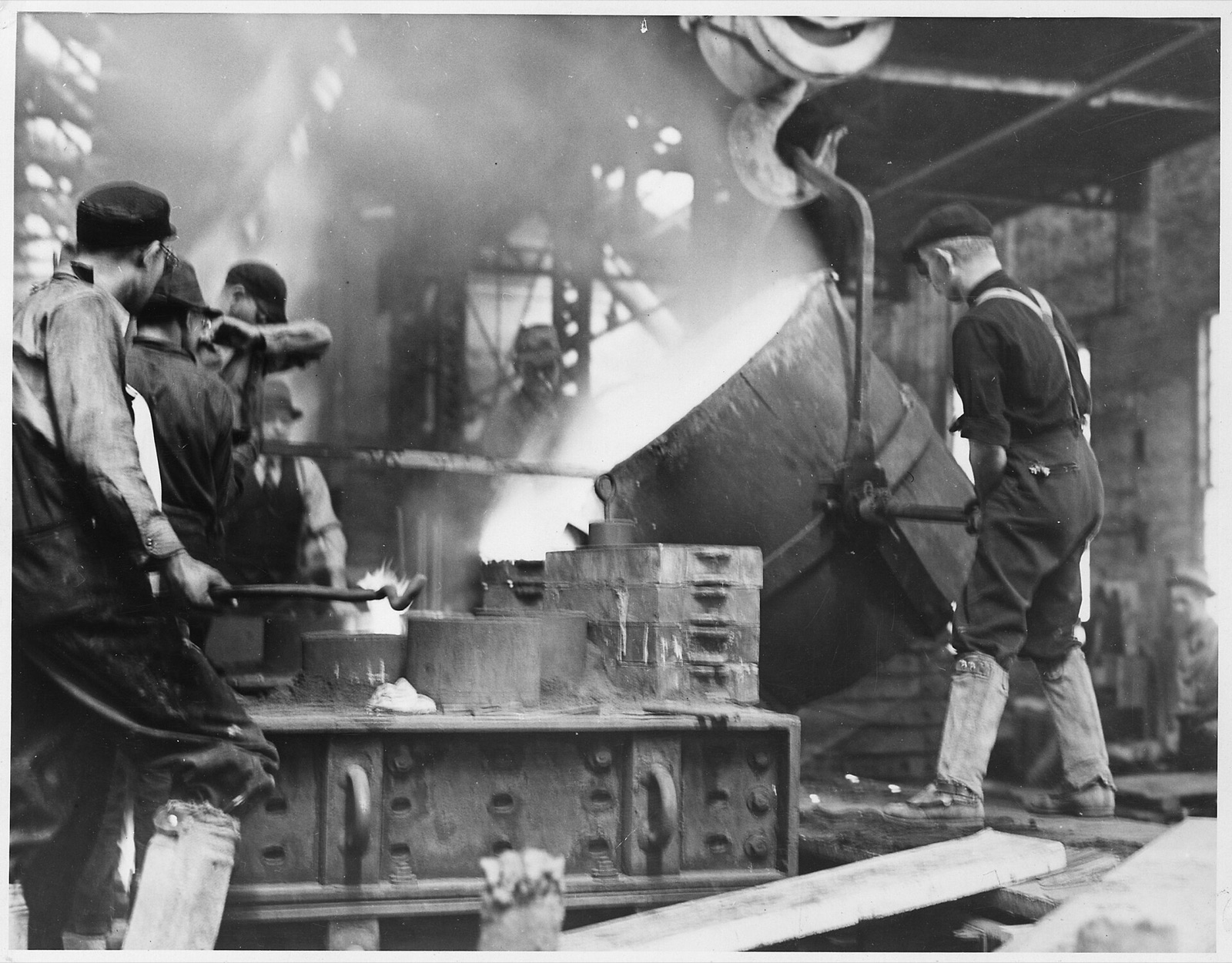 Foundry men in caps watch as a huge crane-held ladle pours glowing metal into stacked molds, one man guiding the ladle wheel