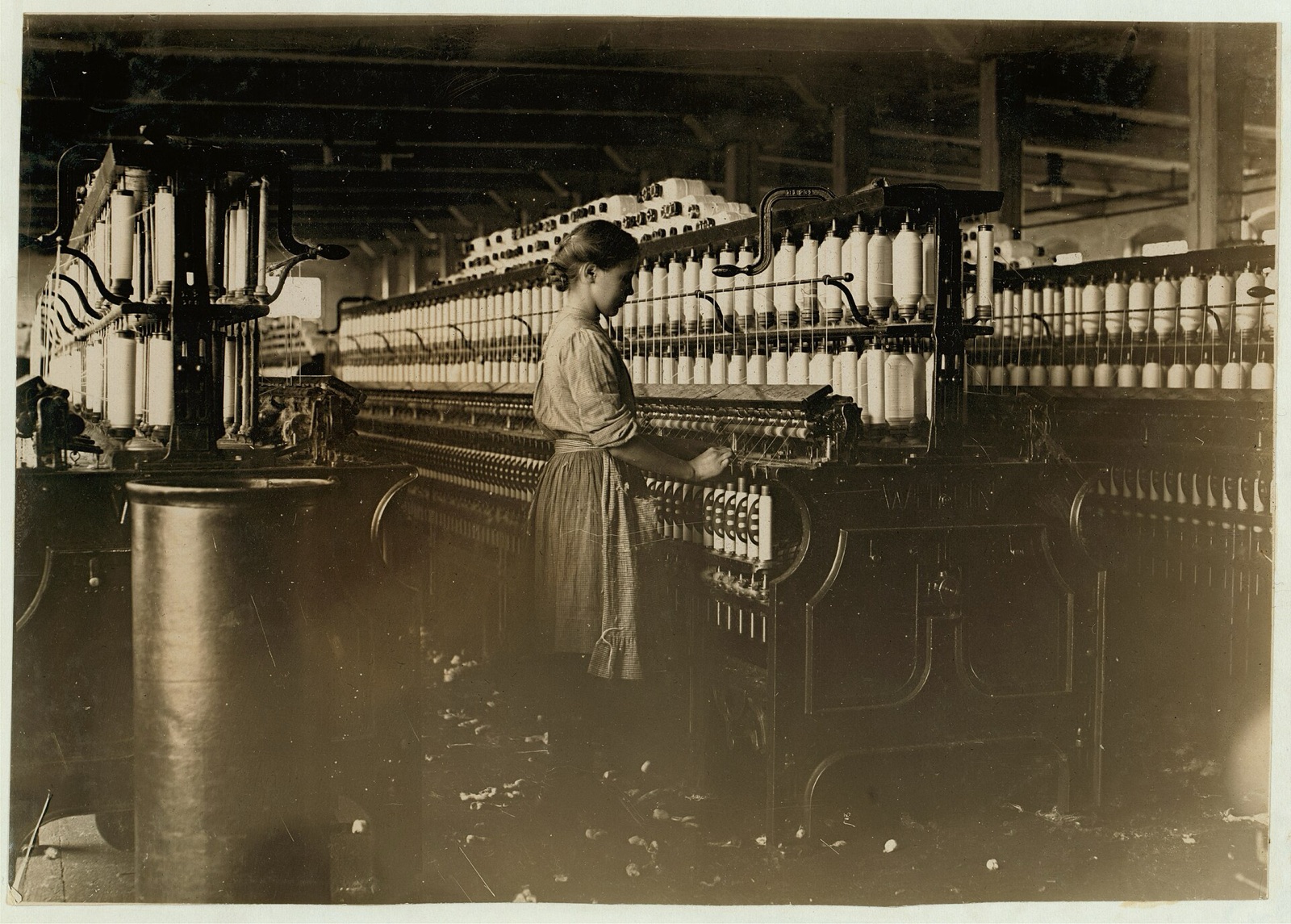 A girl in a pinafore stands at a long spinning frame loaded with spools of cotton yarn, piecing a broken thread