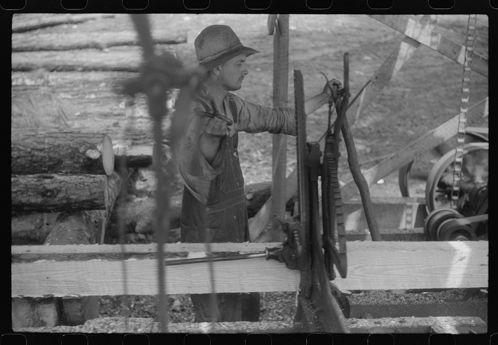 A man in overalls and a felt hat works the lever of a sawmill carriage, with logs stacked behind him and a fresh-cut board in front