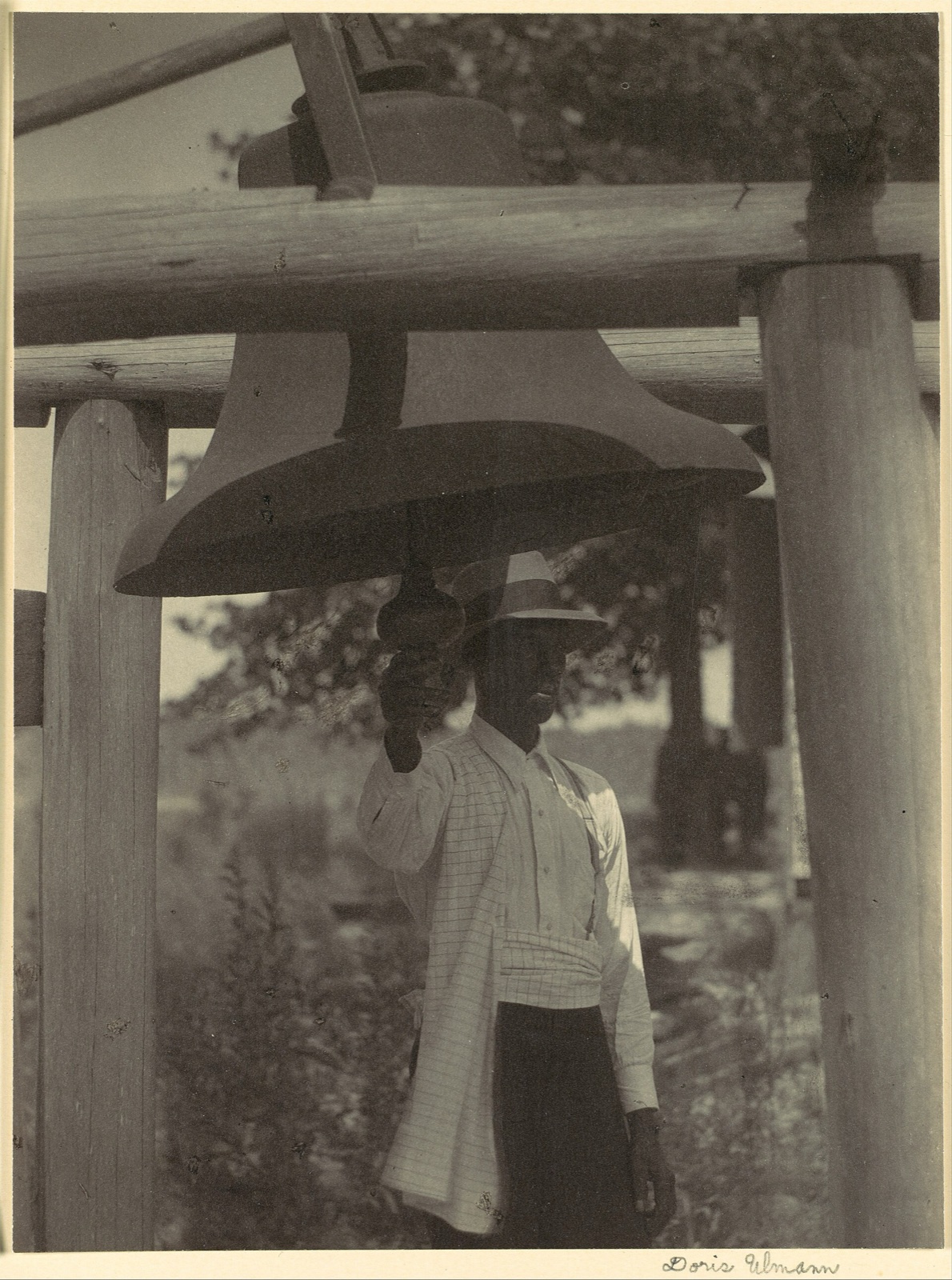 A man in a straw hat and white shirt stands beneath a big bell hung in a timber frame, gripping the bell's clapper
