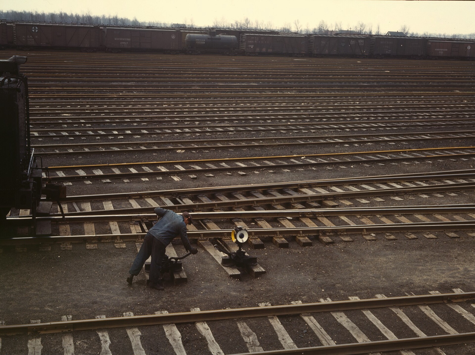 Color photo of a man in a work jacket bending to pull a switch lever beside a switch lamp, with rows of empty tracks and boxcars behind