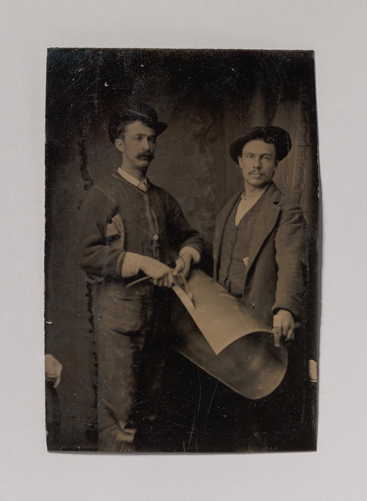 Old tintype of two men in hats holding a large curved sheet of tin, one cutting it with hand shears